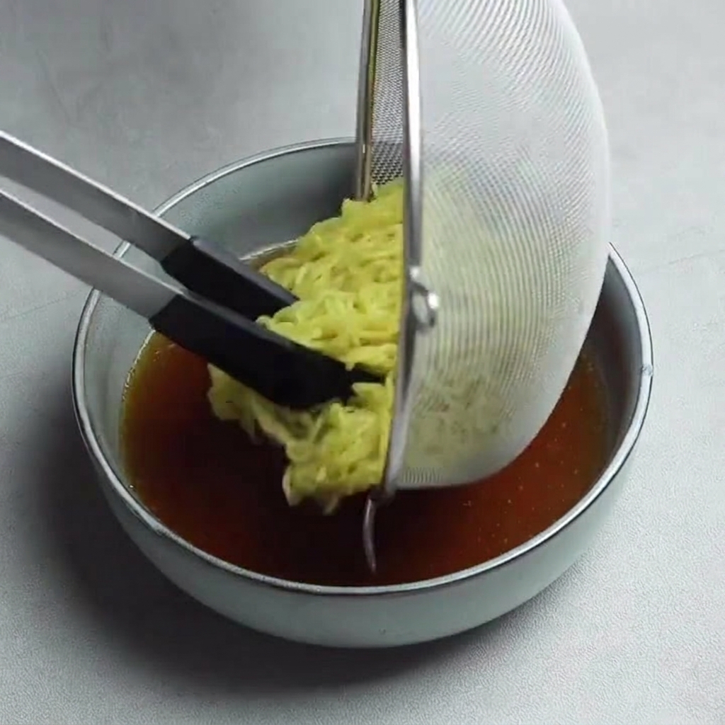 黒いトングを使い、ザルから熱い醤油スープの入った鉢へと黄色い中華麺をそっと移している様子。