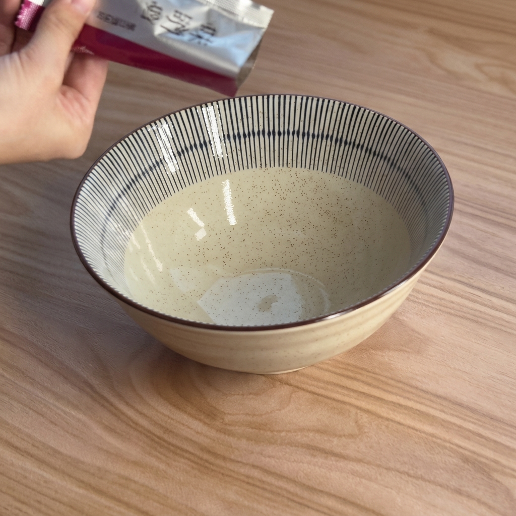 青いストライプの入った白い陶器のラーメン鉢に、シルバーの袋から濃厚な茶色の味噌ペーストを絞り出している様子。