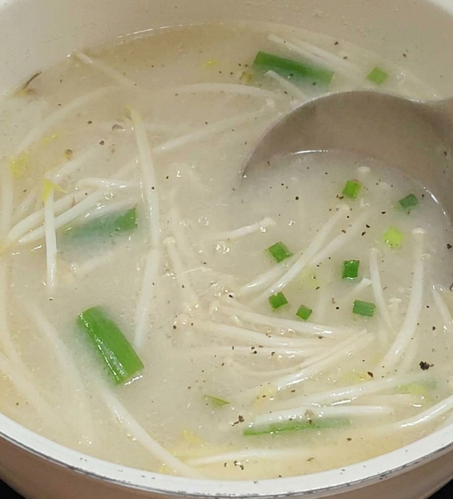 もやしや緑色の野菜が入った鍋に、金属製のおたまから白濁した淡い色のスープが注がれている様子。