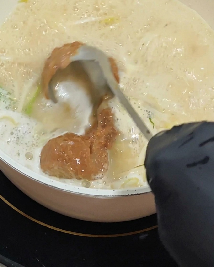 手袋をはめた手がおたまを使い、泡立つクリーミーなスープベースの中に、すくった濃厚な赤茶色の味噌ペーストを混ぜ溶かしている様子。