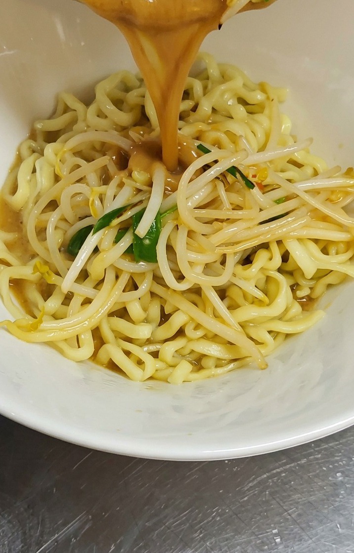 縮れた黄色いラーメンの麺が入った白い丼に、鍋から濃厚な茶色の味噌スープと炒めたもやしが注がれている様子。