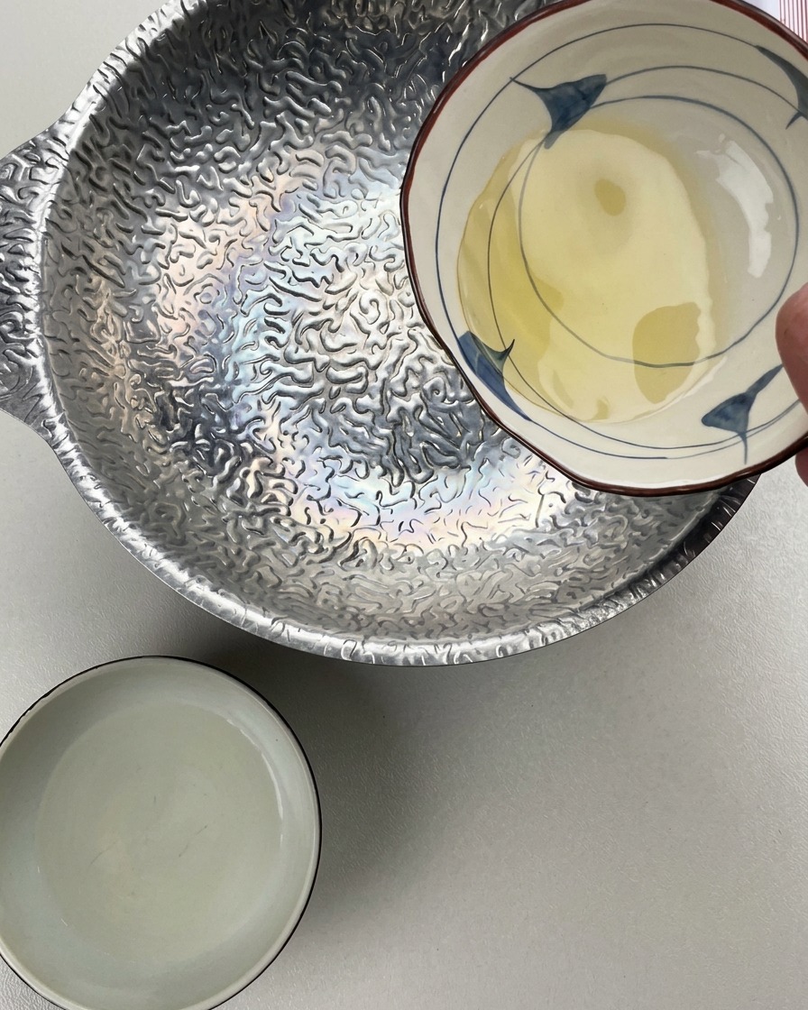 小さな陶器の器から凹凸のある銀色の鍋にみりんを注いでいる様子。横には酒の入った器が置かれている。