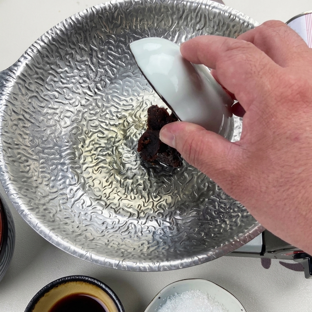 白い小鉢を使って、煮詰まった液体の中に濃い赤味噌をぽってりと落とし入れている手元の様子。