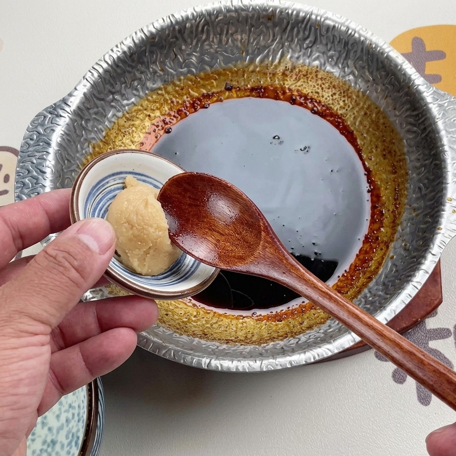 金属製の鍋の中で煮詰まった濃いソースの上に、白っぽい白味噌をすくった木べらをかざしている様子。