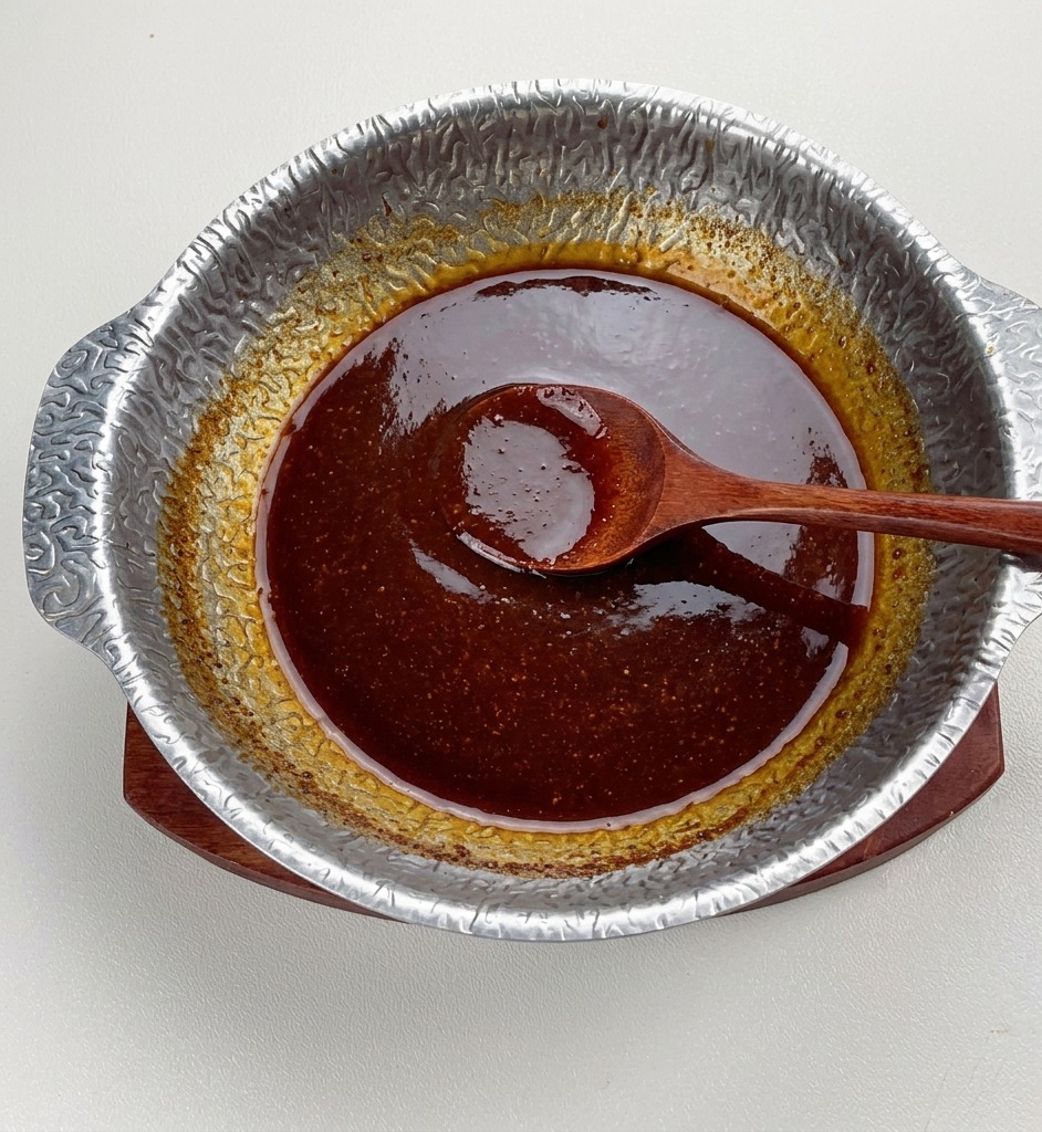 とろみと艶が出て完成した濃い色の味噌つけだれが鍋の中で煮立っており、中央に木べらが置かれている様子。