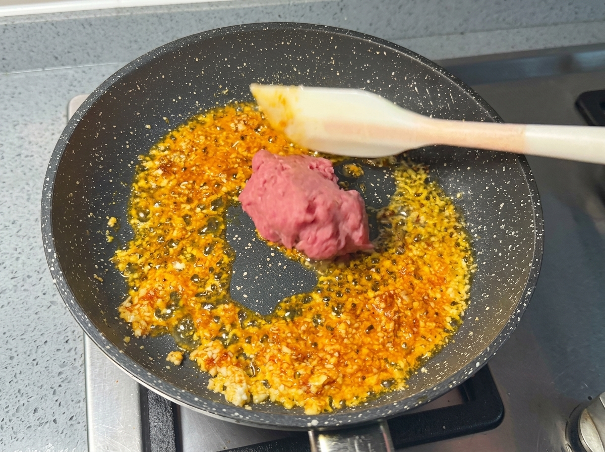 白いシリコンスパチュラを使い、にんにくとエビペーストが香るフライパンに生のピンク色の豚挽き肉を入れている様子。