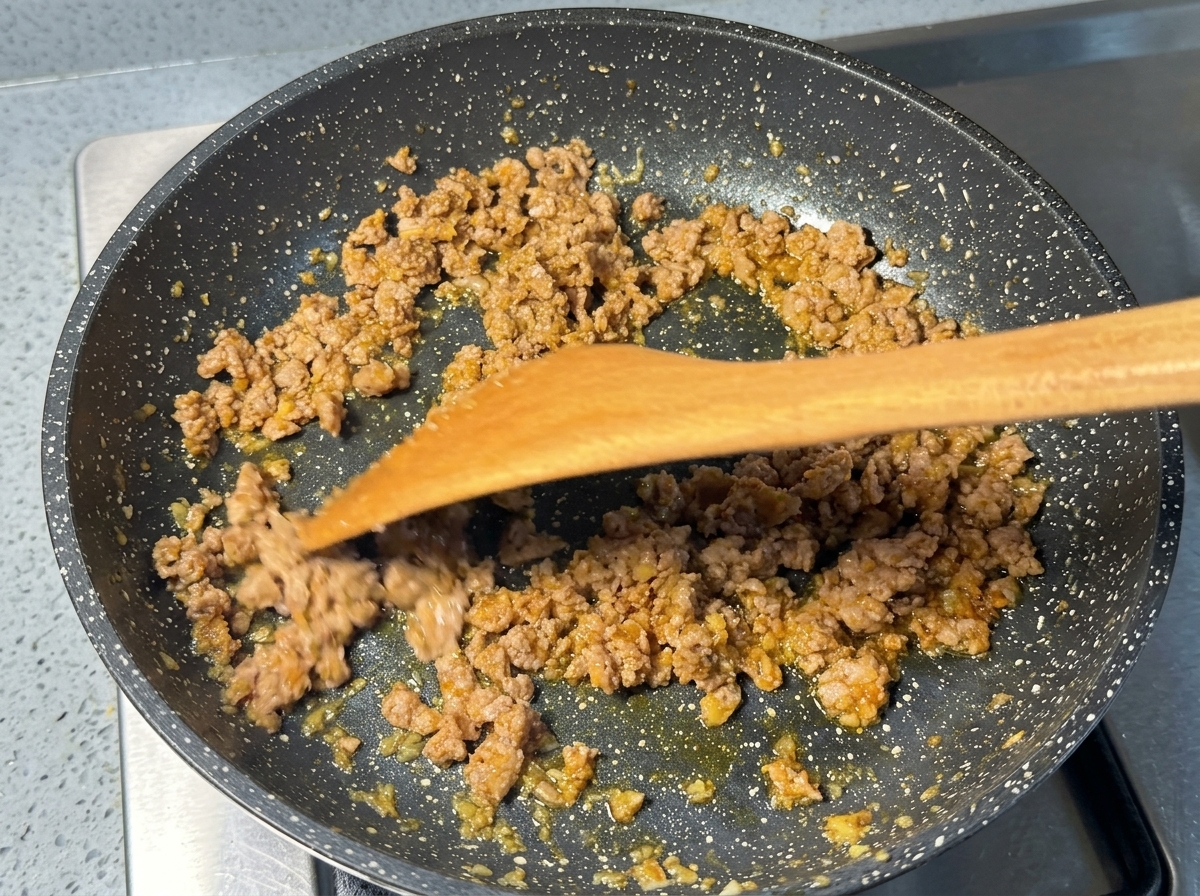 斑点模様のフライパンの中で、木製ヘラを使って炒められているきつね色の豚挽き肉。