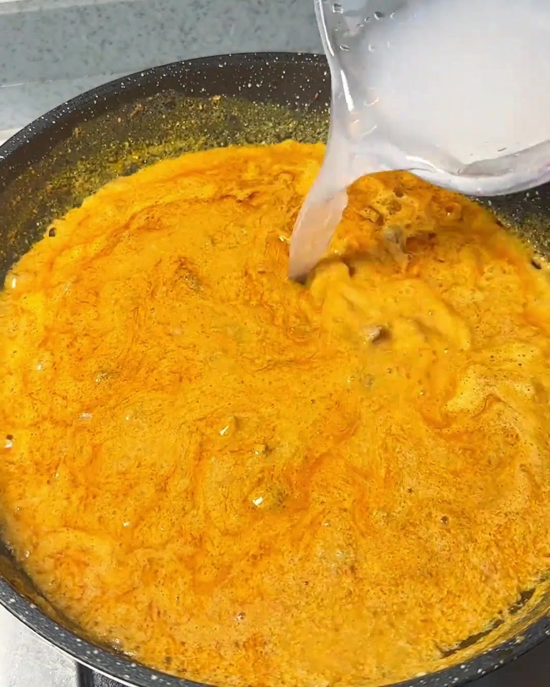 黒いフライパンの中でグツグツと泡立つ濃厚なオレンジ色のカレースープに、容器からきれいな水を注いでいる様子。