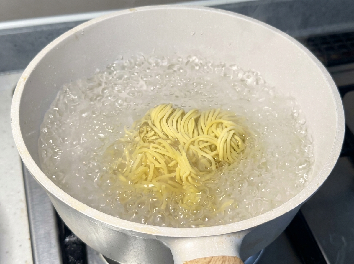 沸騰したお湯が激しく泡立つ白い鍋の中で、黄色い生のラーメンの麺が勢いよく茹でられている様子。
