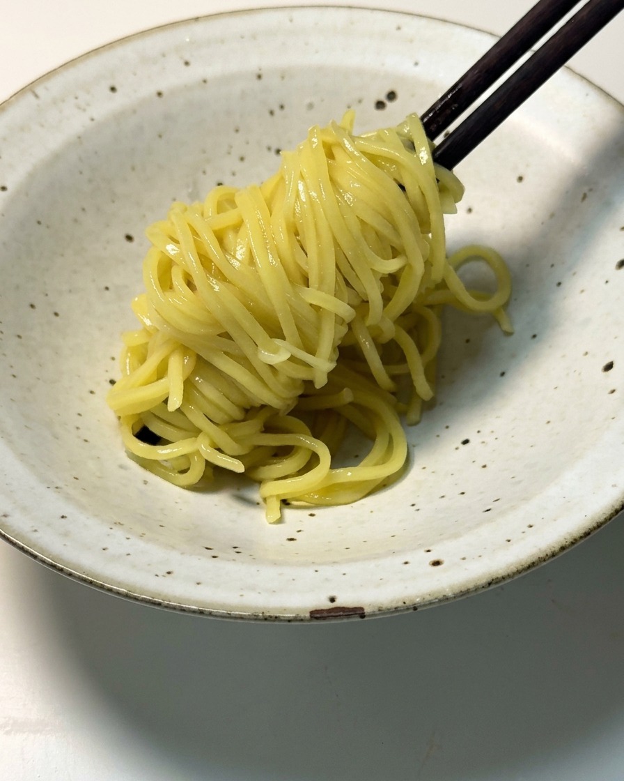 茹で上がった黄色いラーメンの麺を、黒い木箸で持ち上げて斑点模様のある浅い陶器の器に盛り付けている様子。