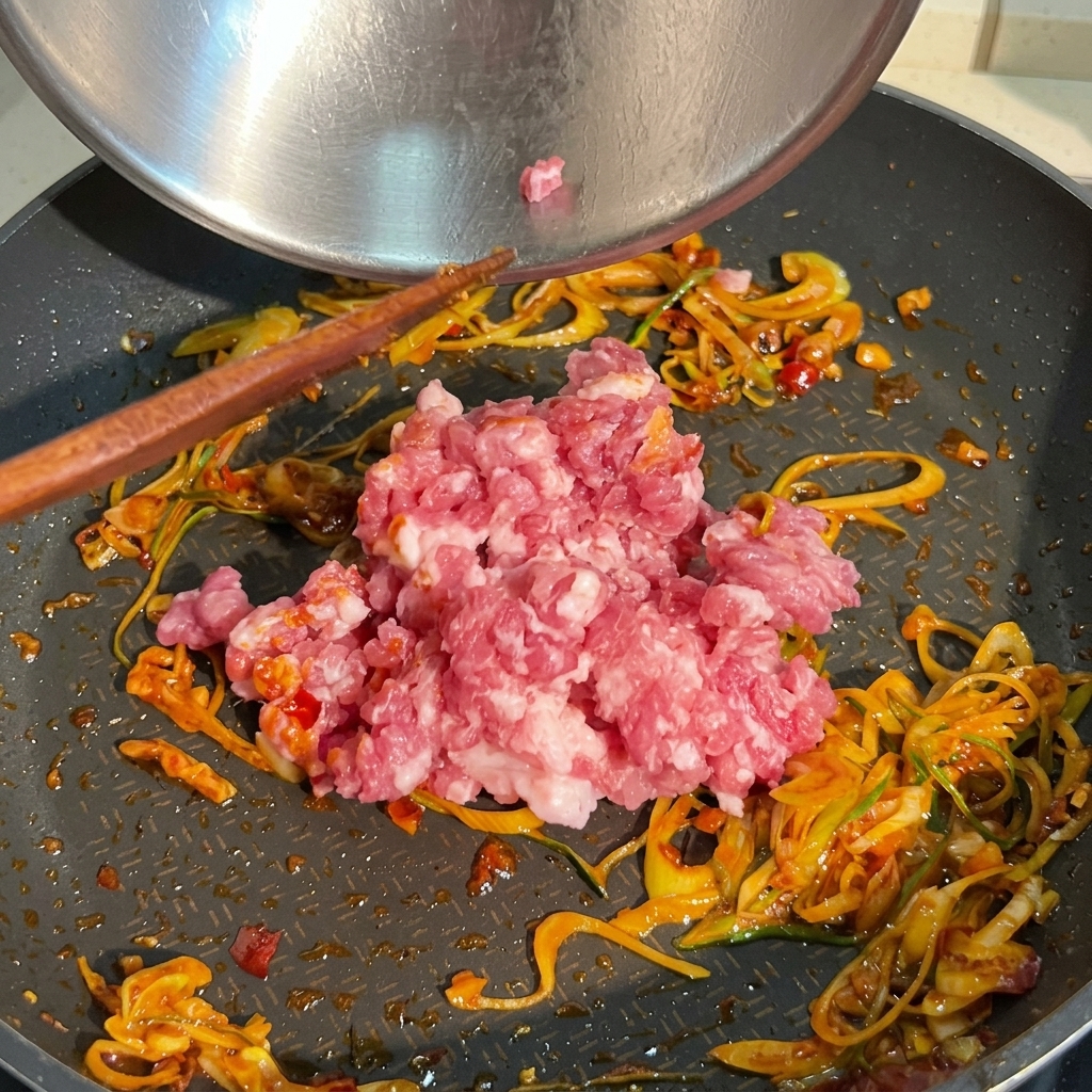 長ねぎと赤色のソースが香るフライパンに、生の豚ひき肉を加える様子。