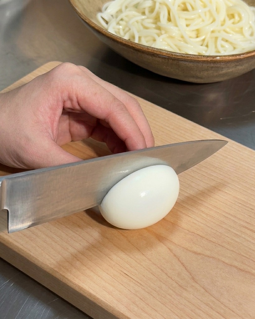 木製のまな板の上できれいな半熟卵を包丁で半分にカットする様子。背景に麺の器が見える。
