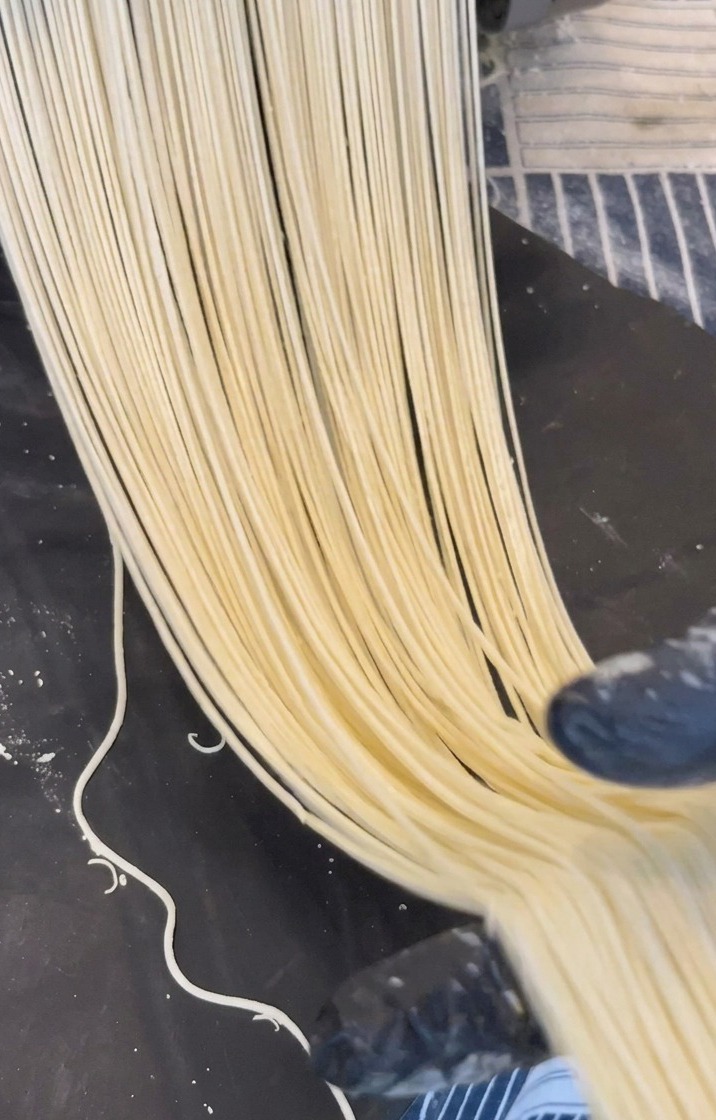 金属製の製麺機から、薄黄色をした細長いラーメンの生麺が黒い台の上に引かれていく様子
