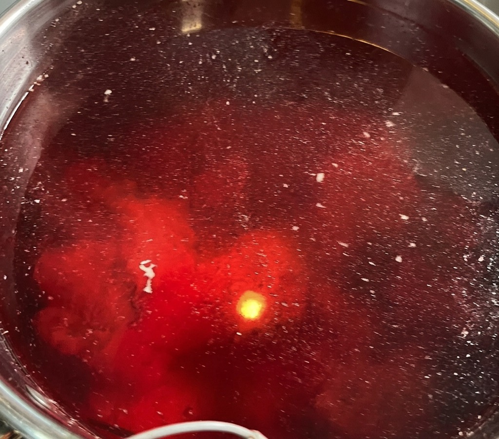 大きなステンレス鍋で豚骨を下茹でしている様子を上から見た写真。水が赤く染まり、表面には白いアクや不純物が浮き出ている。
