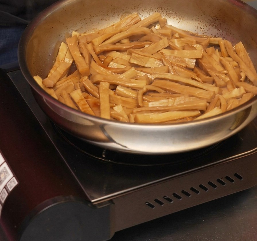 煮汁が煮詰まって濃いタレが絡んだ味付けメンマのフライパンに、ごま油を回し入れて炒める様子。
