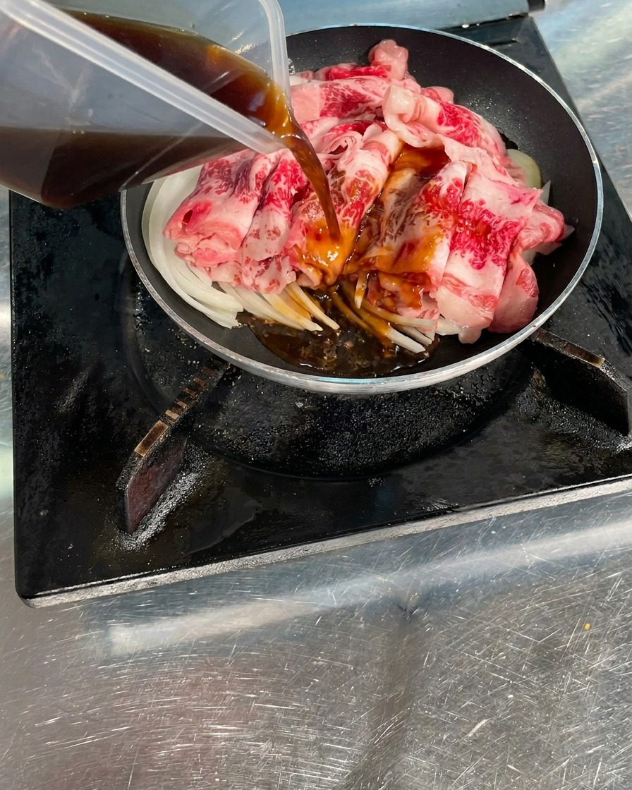 フライパンの中の生肉と玉ねぎの上から、透明な容器に入った濃い茶色のタレを回し入れる様子