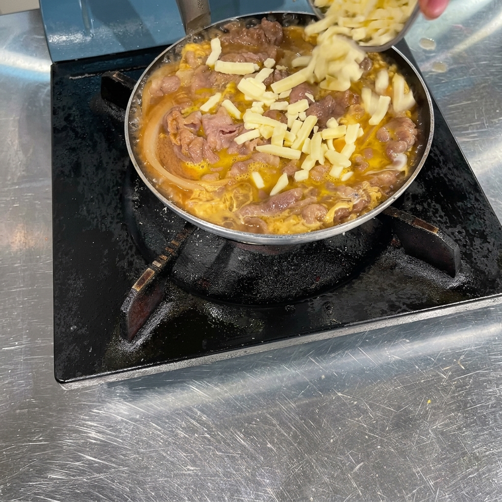 調理された牛肉、玉ねぎ、優しく固まった卵が入ったフライパンに、手でピザ用チーズを散らしている様子