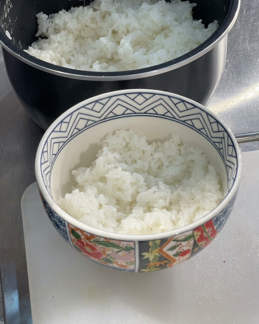 炊飯器の横のまな板の上に置かれた、炊きたてでふっくらとした白米が半分ほど盛られた陶器の丼ぶり