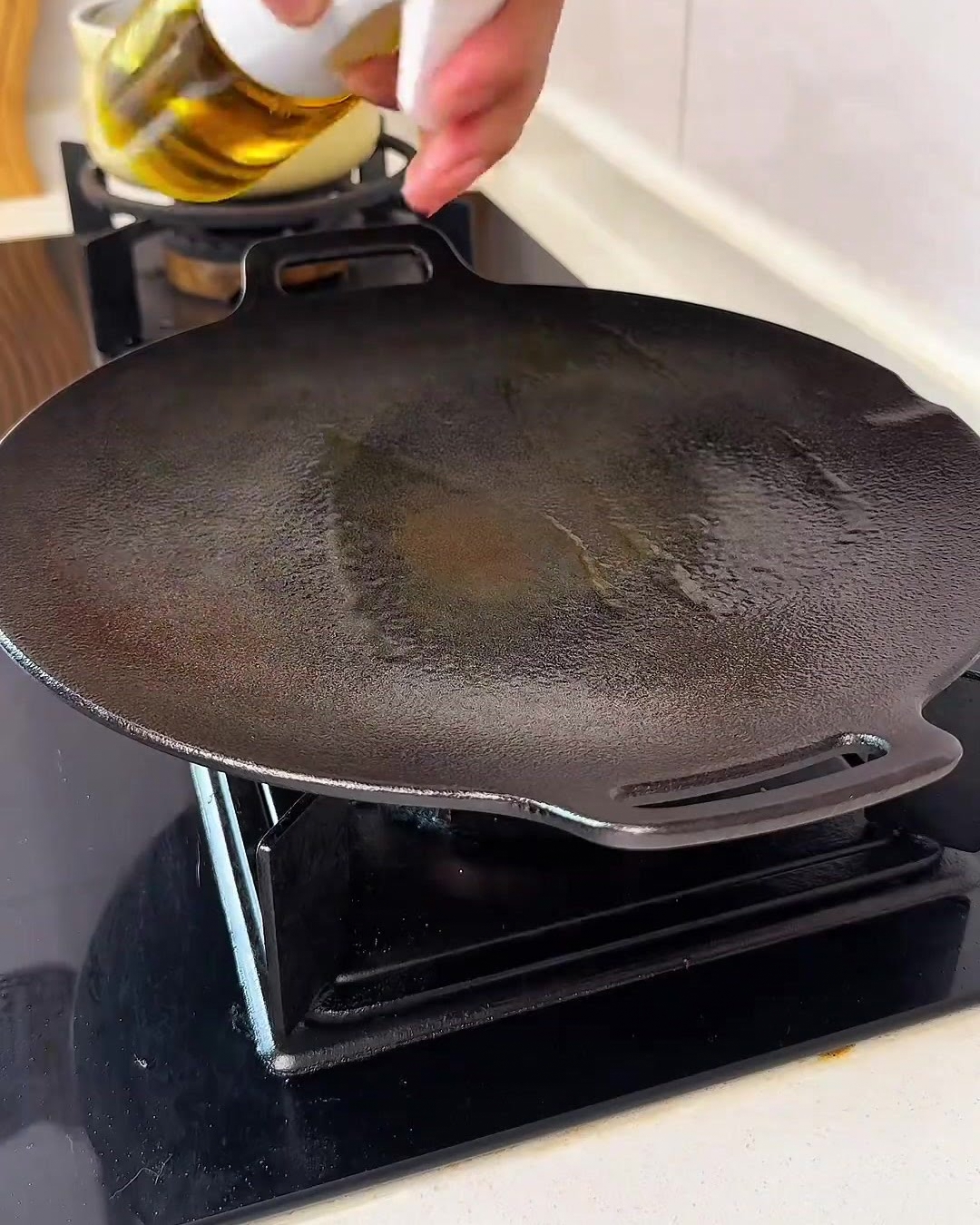 コンロの上の黒い丸型の鉄板に、スプレーで黄金色のサラダ油を細かく吹き付けている様子。