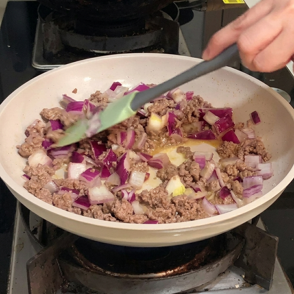 フライパンの中で、細かく刻まれた紫と白の赤玉ねぎが、色づいた牛ひき肉と一緒に炒められている様子。