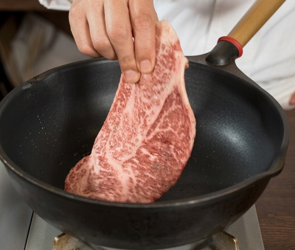 黒く熱したスキレットに、黒こしょうで下味をつけた大きな生の霜降りステーキ肉を手で丁寧に入れている様子。