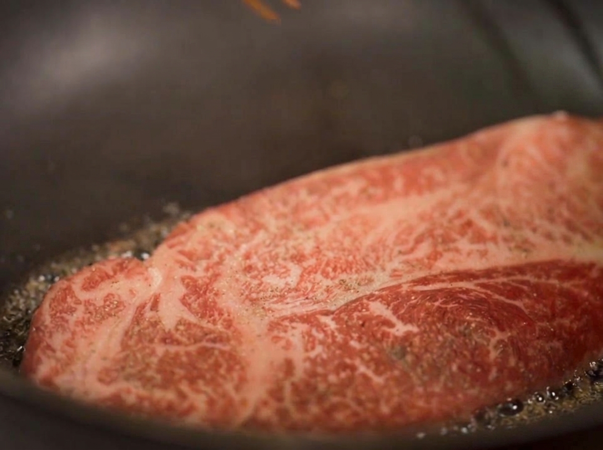 厚切りのステーキが油で焼かれているクローズアップ写真。湯気が立ち上り、底の縁が黄金色に変わっている様子。