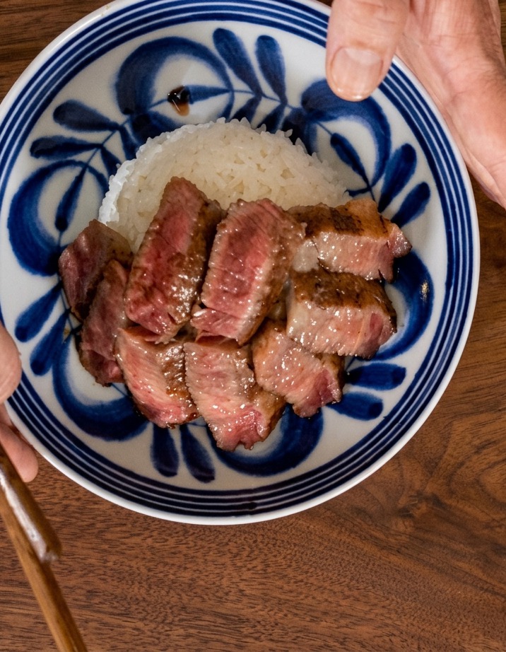 青い模様の入った丼を片手で持ち、もう片方の手で白ご飯の上にステーキのスライスを放射状に重ねて並べている俯瞰写真。