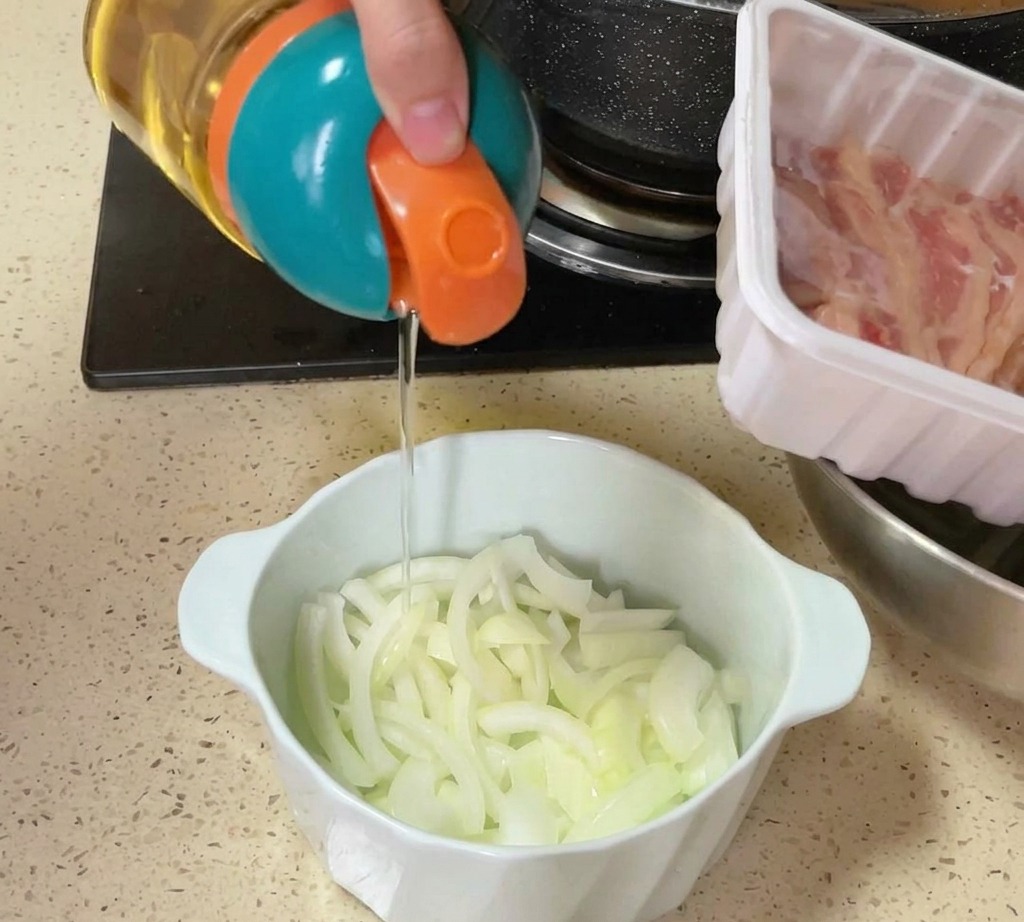 キッチンの調理台の上で、薄切りにした生の玉ねぎが入ったボウルに、ハンディディスペンサーから油や塩を振りかけている様子。