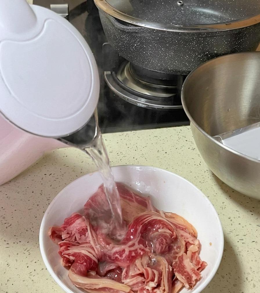 ピンクと白の電気ケトルから、ボウルに入った生の牛バラ薄切り肉に沸騰したお湯を注いでいる様子。