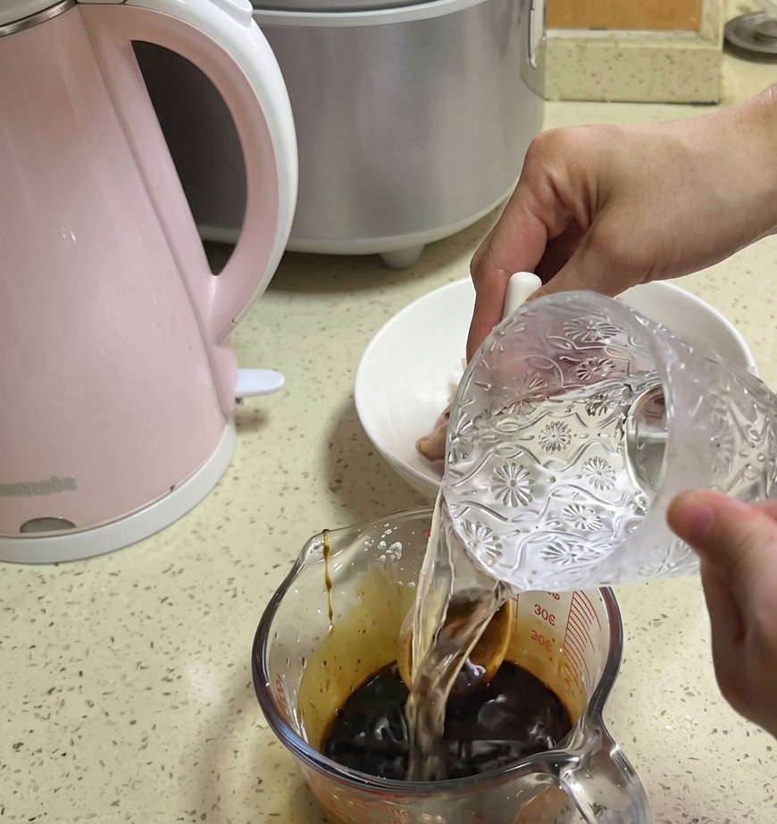 醤油ベースの濃い茶色のタレが入った計量カップに、ガラスのピッチャーからきれいな水を注いでいる様子。