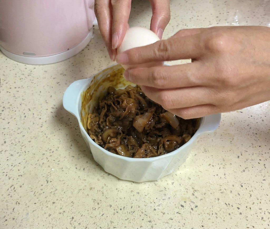 黄色いスライスチーズが2枚乗った牛肉のボウルの上で、両手を使って白い卵を割り入れている様子。