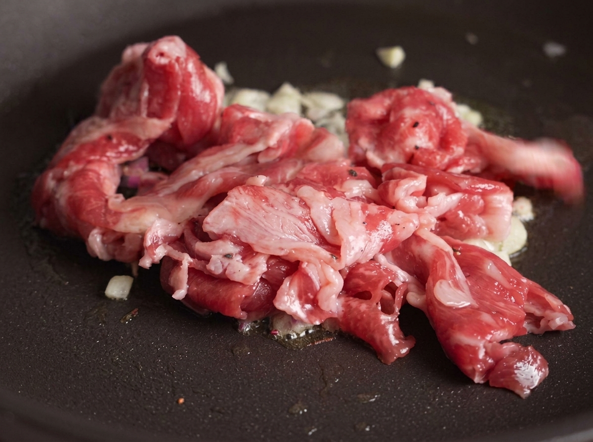 にんにくの香りが移ったフライパンに生の牛カルビ肉を投入し、熱で肉の一部分が茶色く変わり始めている様子。