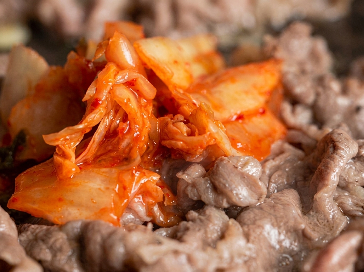 フライパンの中で火が通りかけた薄切り牛肉の中に、鮮やかな赤色のキムチをたっぷりと投入して混ぜ合わせているカット。