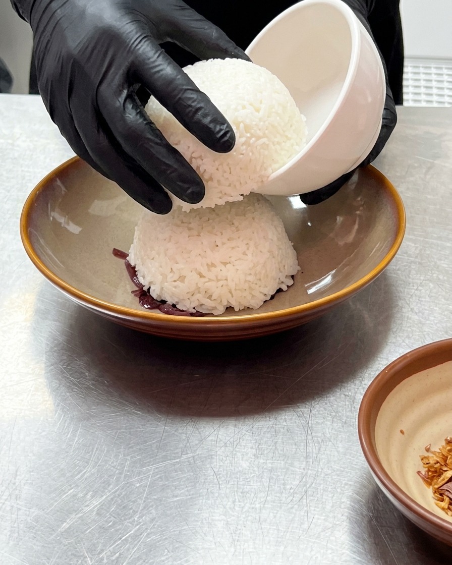 手袋を着用した手が小さな白いお椀を持ち上げ、紫玉ねぎの上にきれいに型取られたドーム状のご飯が現れる様子。