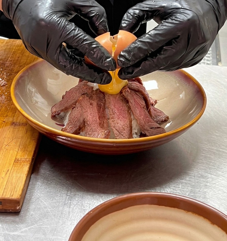 手袋を着用した両手で、山のように盛られた牛肉の頂上にあるへこみに、温泉卵を優しく割り落としている様子。