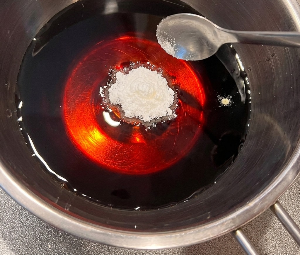 ステンレス製の鍋に入った濃い醤油ベースの液体に、スプーン1杯の白いだしの素を加えている様子。