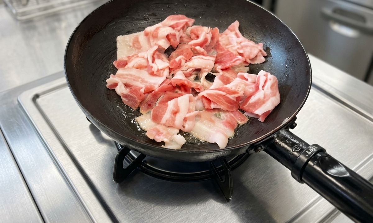 ステンレス製のコンロの上で、黒いフライパンを使って豚バラ肉のスライスを炒めている様子。