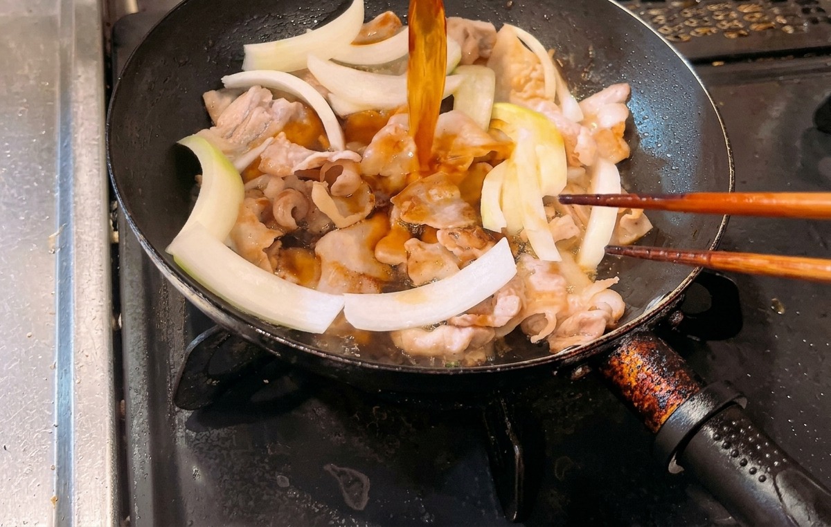 炒めた豚バラ肉と生の玉ねぎのスライスが入ったフライパンに、琥珀色の合わせタレを注ぎ入れている様子。