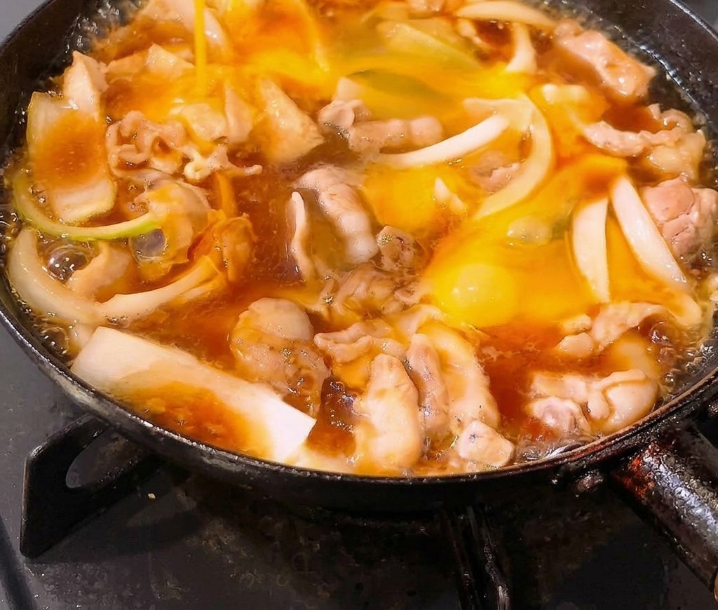 出汁ベースのタレで煮込まれている豚バラ肉と玉ねぎのフライパンに、溶き卵を注ぎ入れている様子。