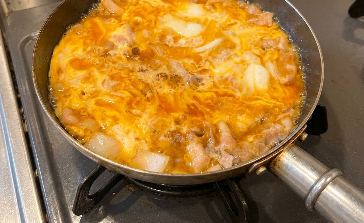 コンロの上で煮込まれる他人丼のフライパン。豚肉や玉ねぎの周りで黄色い卵が半熟状に固まってきている様子。