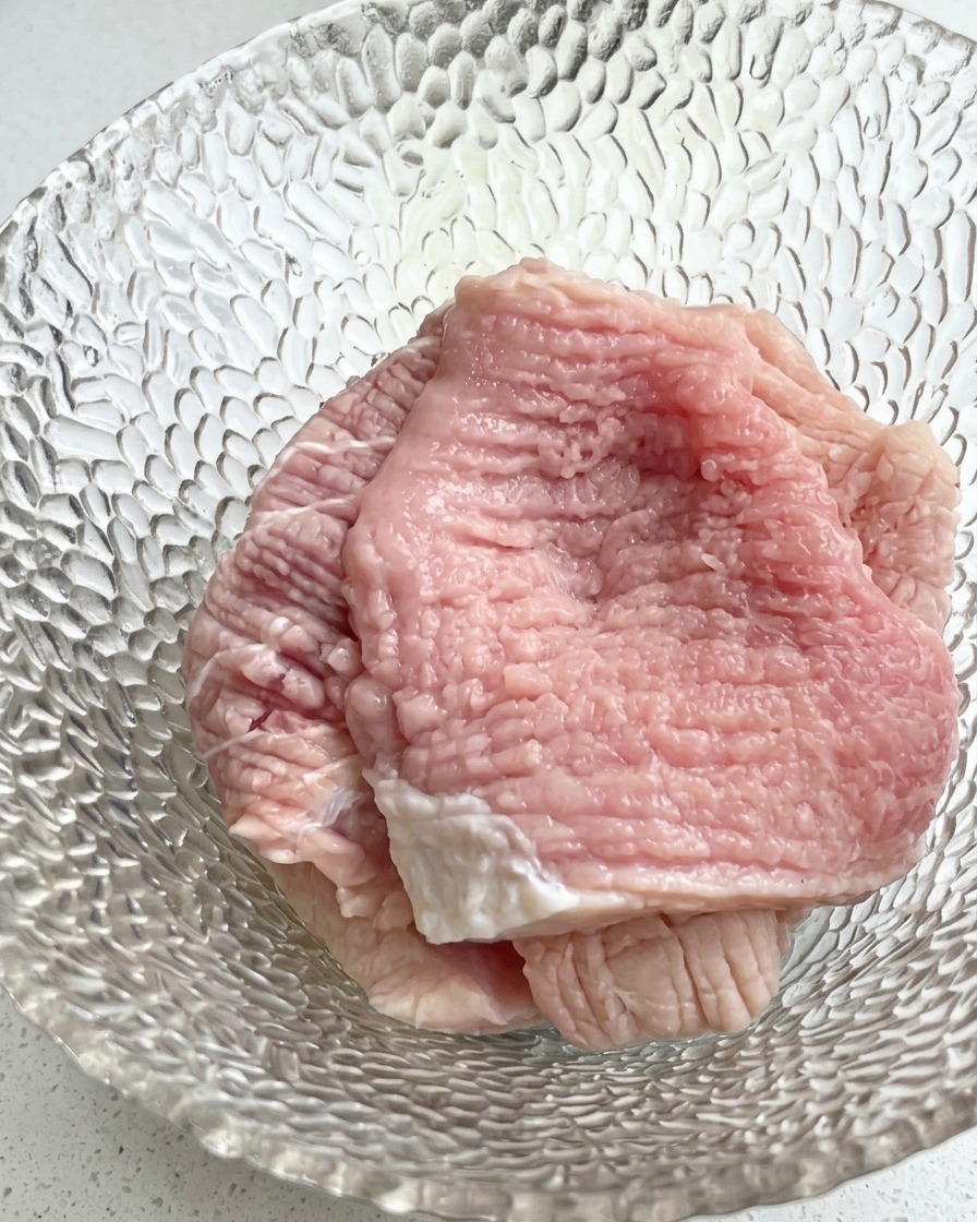 ガラスのボウルに入った生の豚肉スライスの上に、刻んだ青ねぎと調味料が加えられているクローズアップ画像。