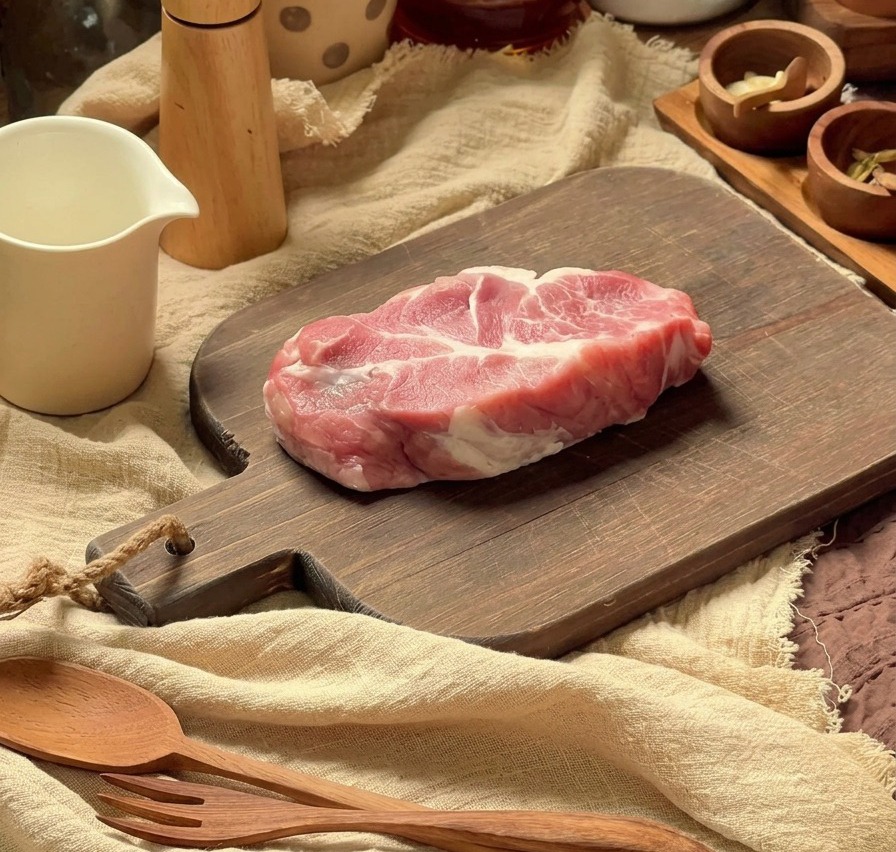 木製のまな板の上で調理器具を背景に、下ごしらえされる生の豚肩ロース肉。