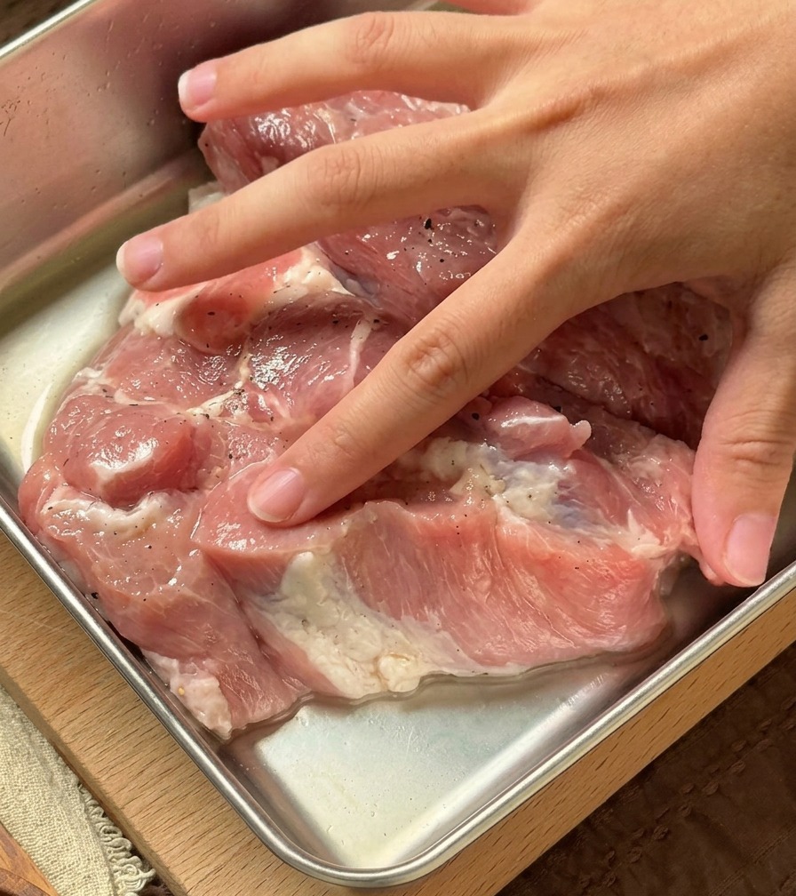 金属製のバットの中で、生の豚肉に透明な香味液を手でもみ込んでいる様子。