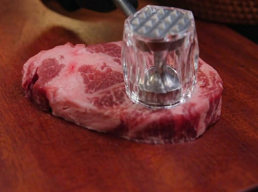 木製のまな板の上で、金属製の肉叩きを使って生の豚肩ロース肉を丁寧に叩いている様子。