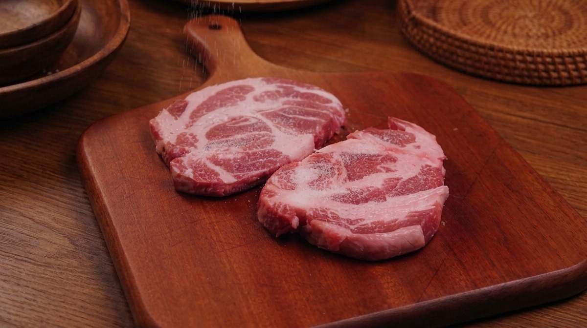 まな板の上に並べられた生豚肩ロース肉に、手で塩と黒こしょうをバランスよく振っている様子。