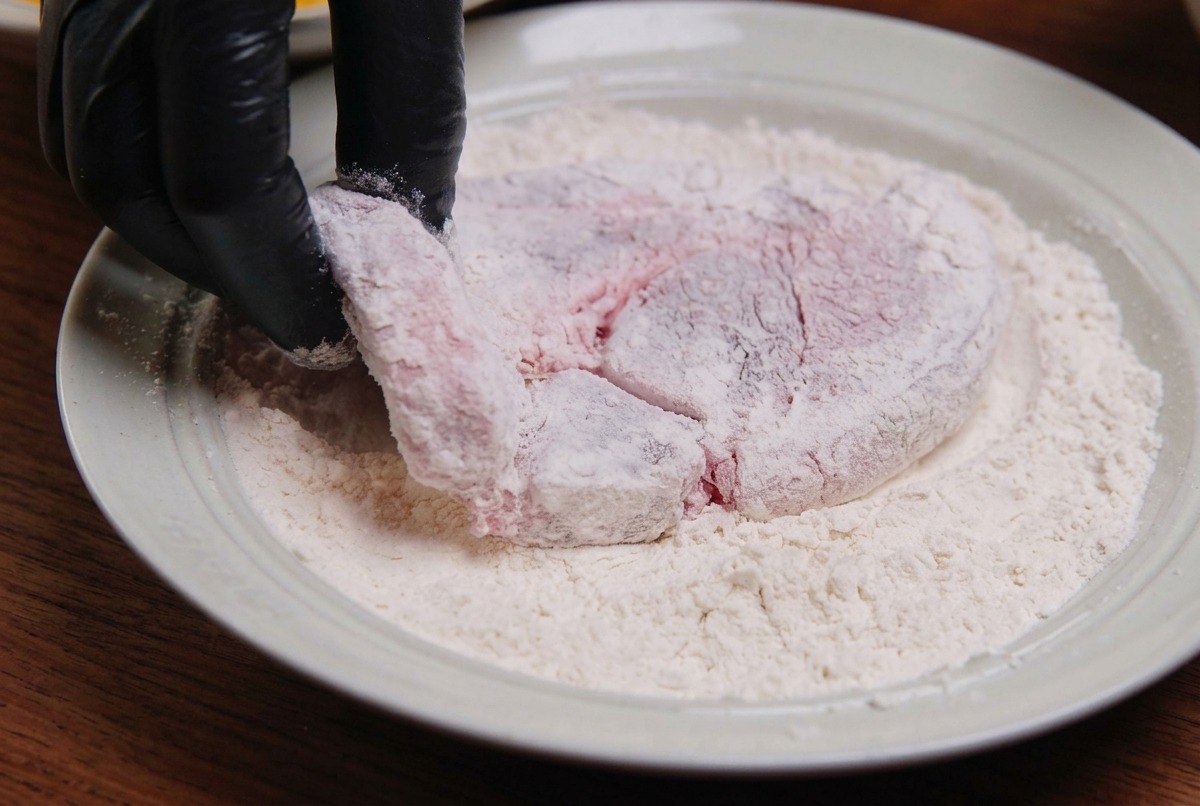 浅い皿の中で、生の豚肉に小麦粉を薄く均一にまぶしている様子。