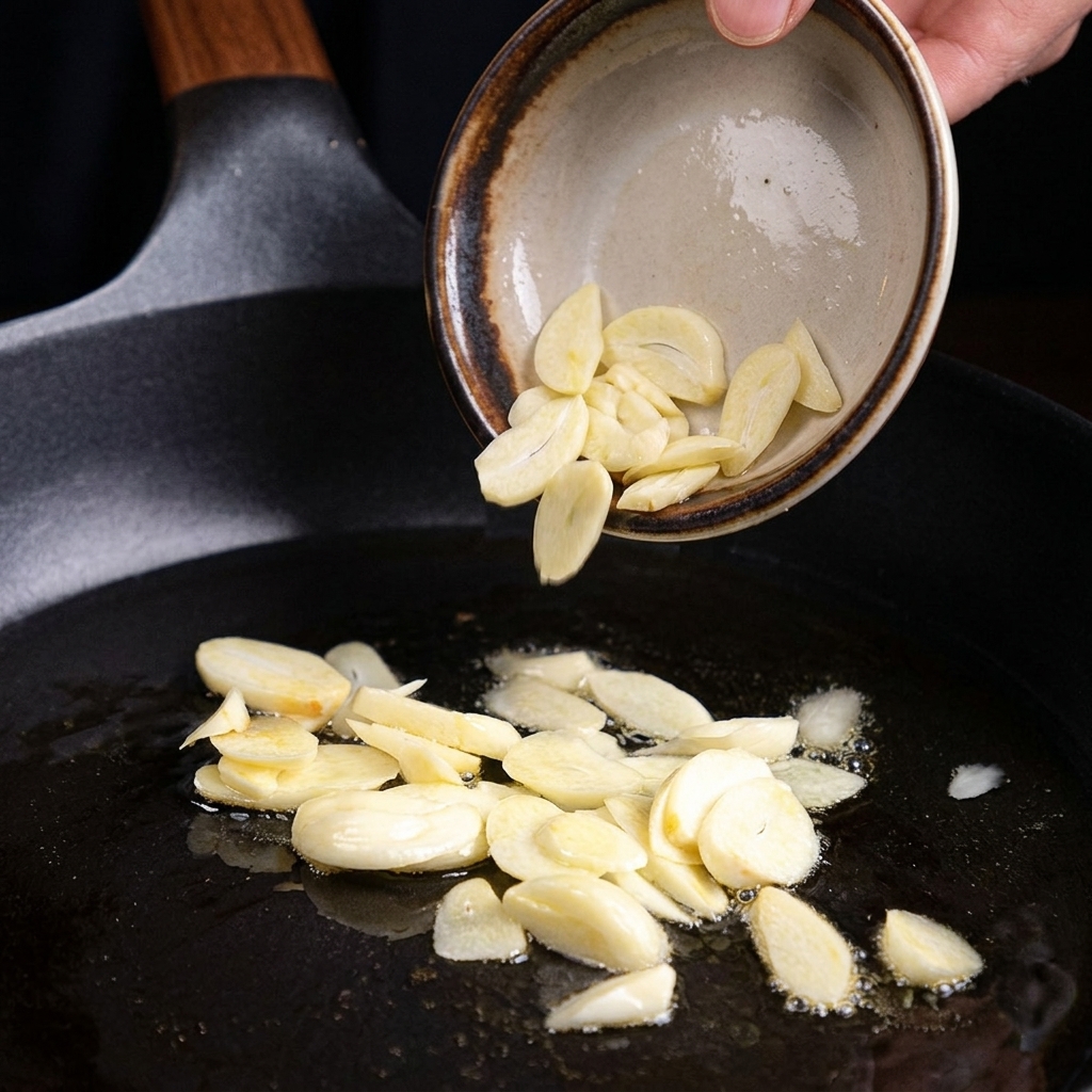 油を熱したフライパンに、小さなボウルからスライスした生にんにくを滑らせるように入れて揚げ焼きにしている様子。