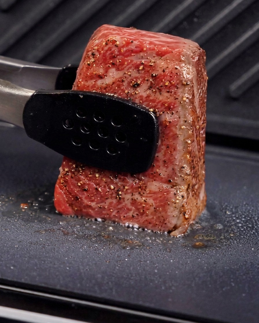 グリルプレートの上で、トングを使って厚切りの肉を垂直に立て、側面の脂身を焼いている様子。湯気と油がジューシーに弾けている。