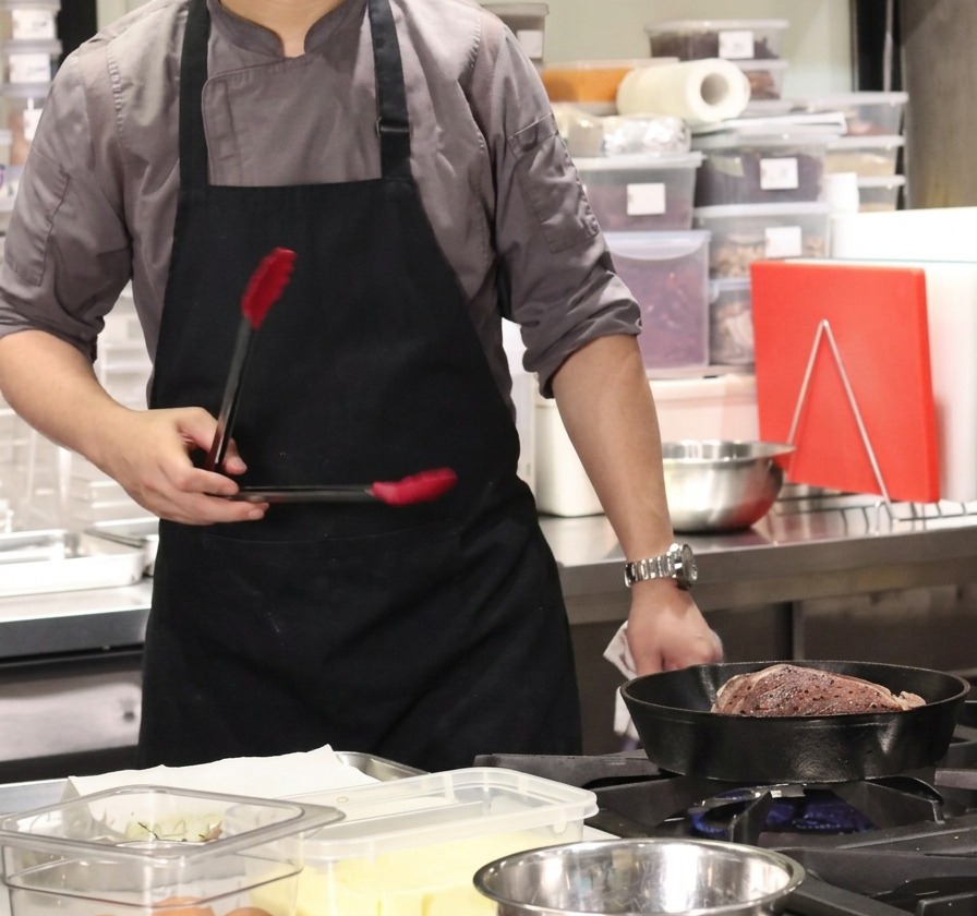 熱した鋳鉄製のフライパンで、トングを使い大きな牛塊肉の脂身の面をしっかりと焼くシェフの手元