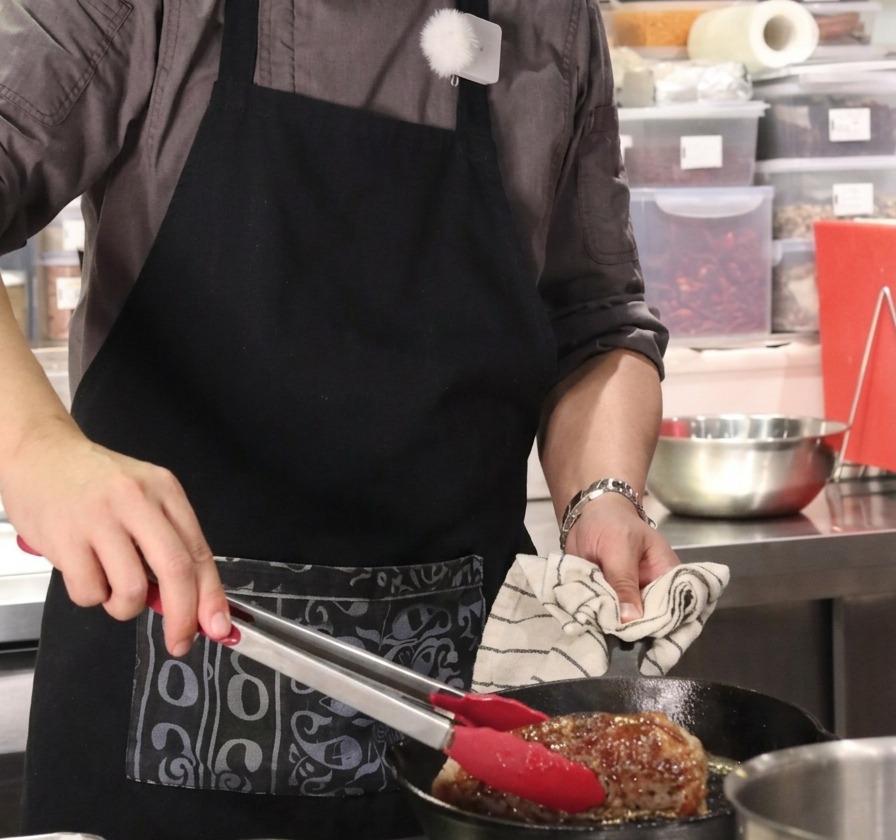 赤いグリップのトングで大きな牛塊肉を垂直に立て、鋳鉄製のフライパンで全面を焼くクローズアップ
